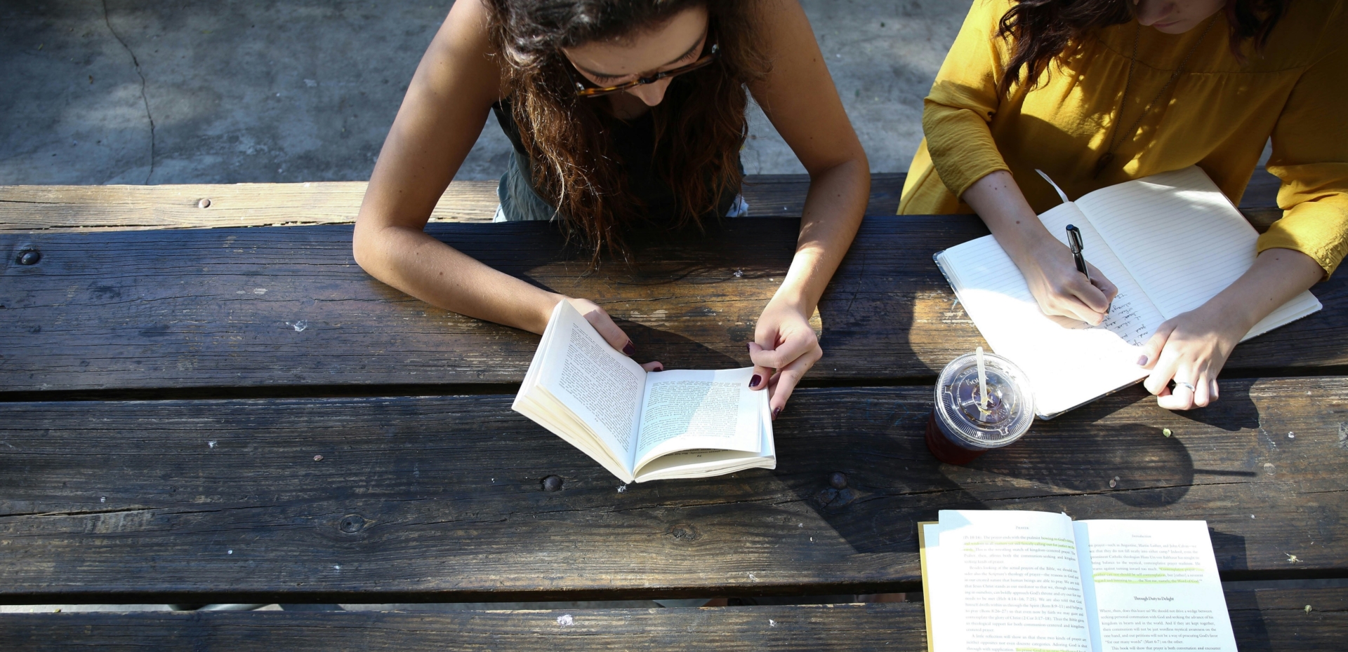 Three friends read books outdoors while seated at a picnic table.