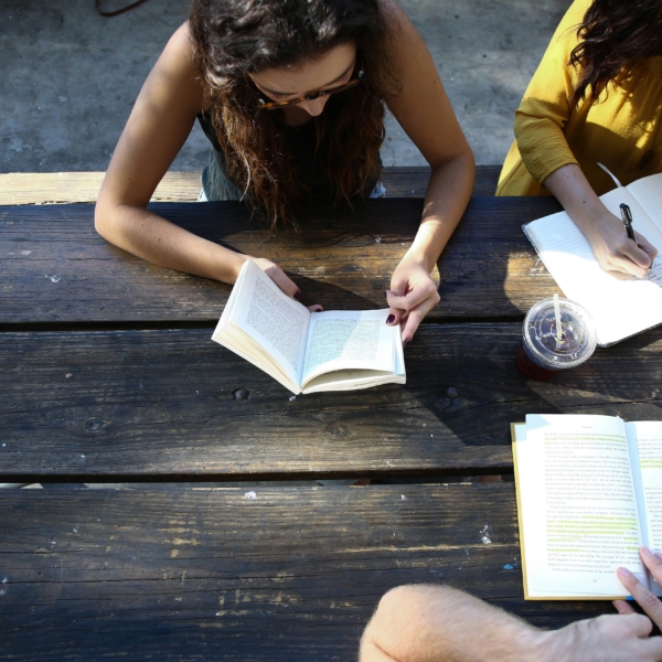 Three friends read books outdoors while seated at a picnic table.