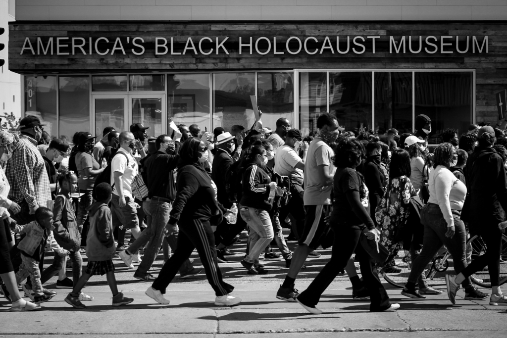 A group of protestors, about six people deep marches calmly but purposefully on the sidewalk directly outside America's Holocaust Museum in a flat, well-composed black-and-white photograph.
