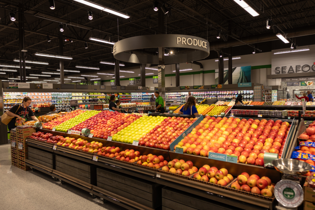 Shoppers select their fruit among rows of produce while a green-uniformed Publix employee restocks.